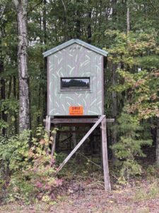 Camouflage-painted wooden box blind on legs at the edge of the woods